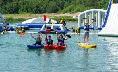 Huge inflatable obstacle course in middle of lake, with people canoing and paddle boarding nearby